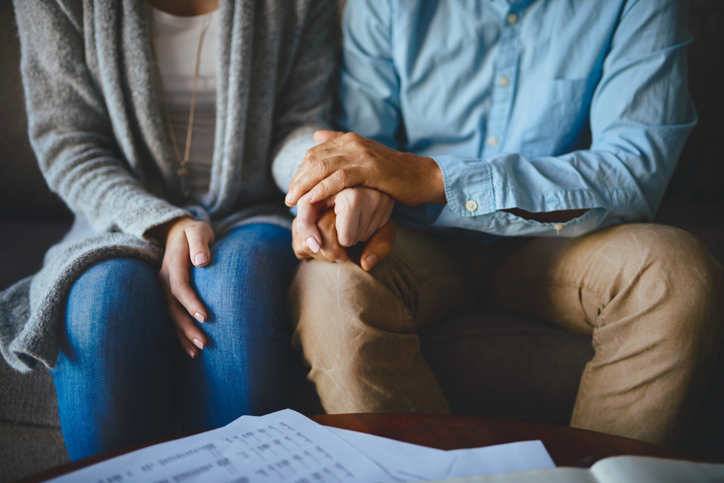 A photo of a couple sitting on a couch holding hands.